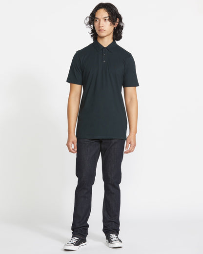A person with long, curly dark hair stands facing forward in a Volcom Men's Wowzer Polo Short Sleeve Shirt, paired with dark jeans and black low-top sneakers against a plain white background.