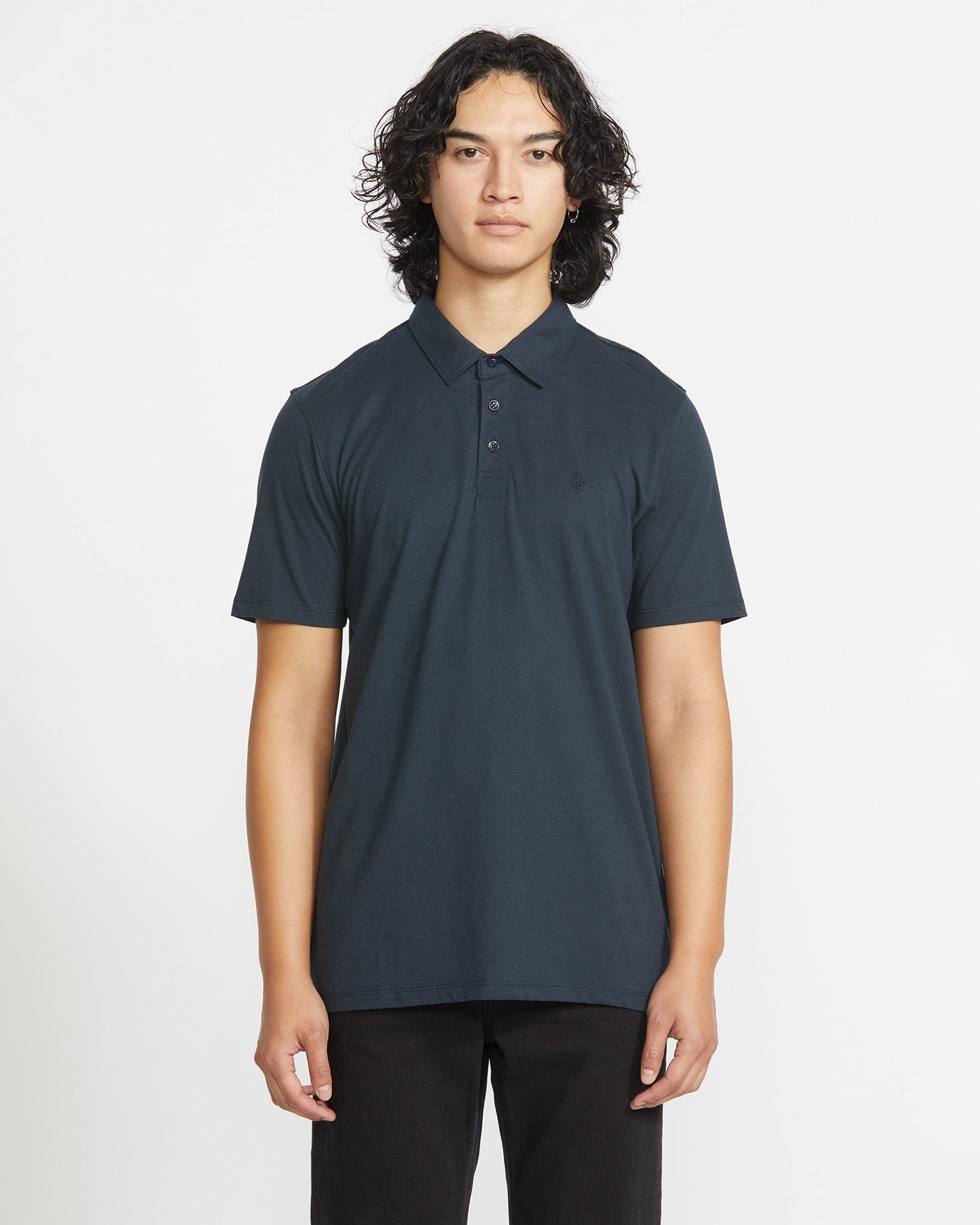 A person with long, curly dark hair faces forward, wearing the Volcom Men's Wowzer Polo Short Sleeve Shirt and black pants against a plain white background.