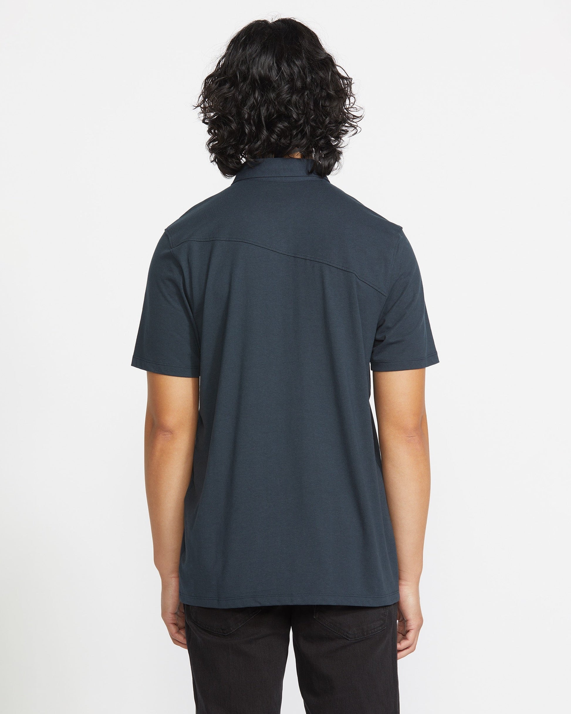 A person with shoulder-length curly hair is shown from the back, wearing the Volcom Men's Wowzer Polo Short Sleeve Shirt and dark pants, standing against a plain white background.