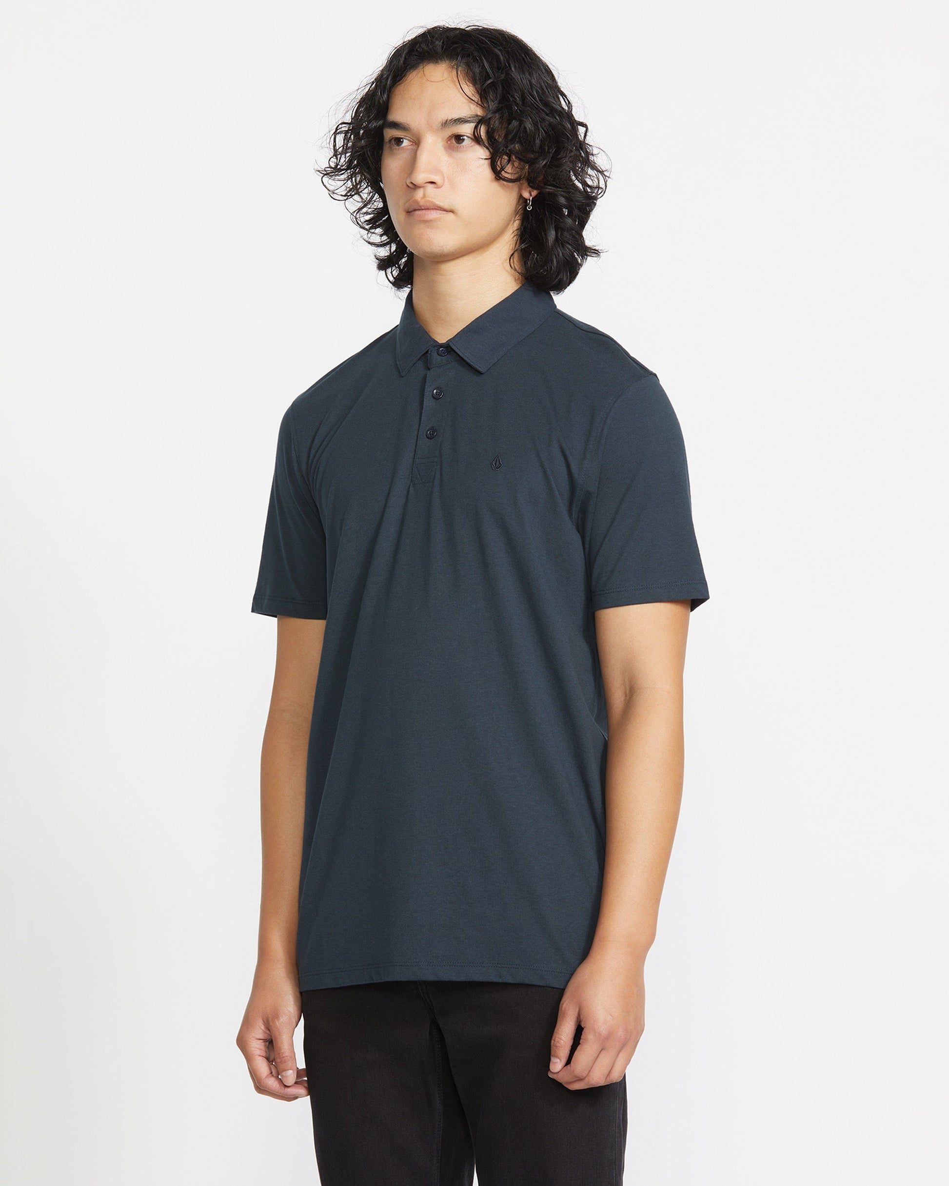 A young man with wavy, shoulder-length dark hair wears the Volcom Men's Wowzer Polo Short Sleeve Shirt, standing against a plain white background and looking slightly to the side with a neutral expression.