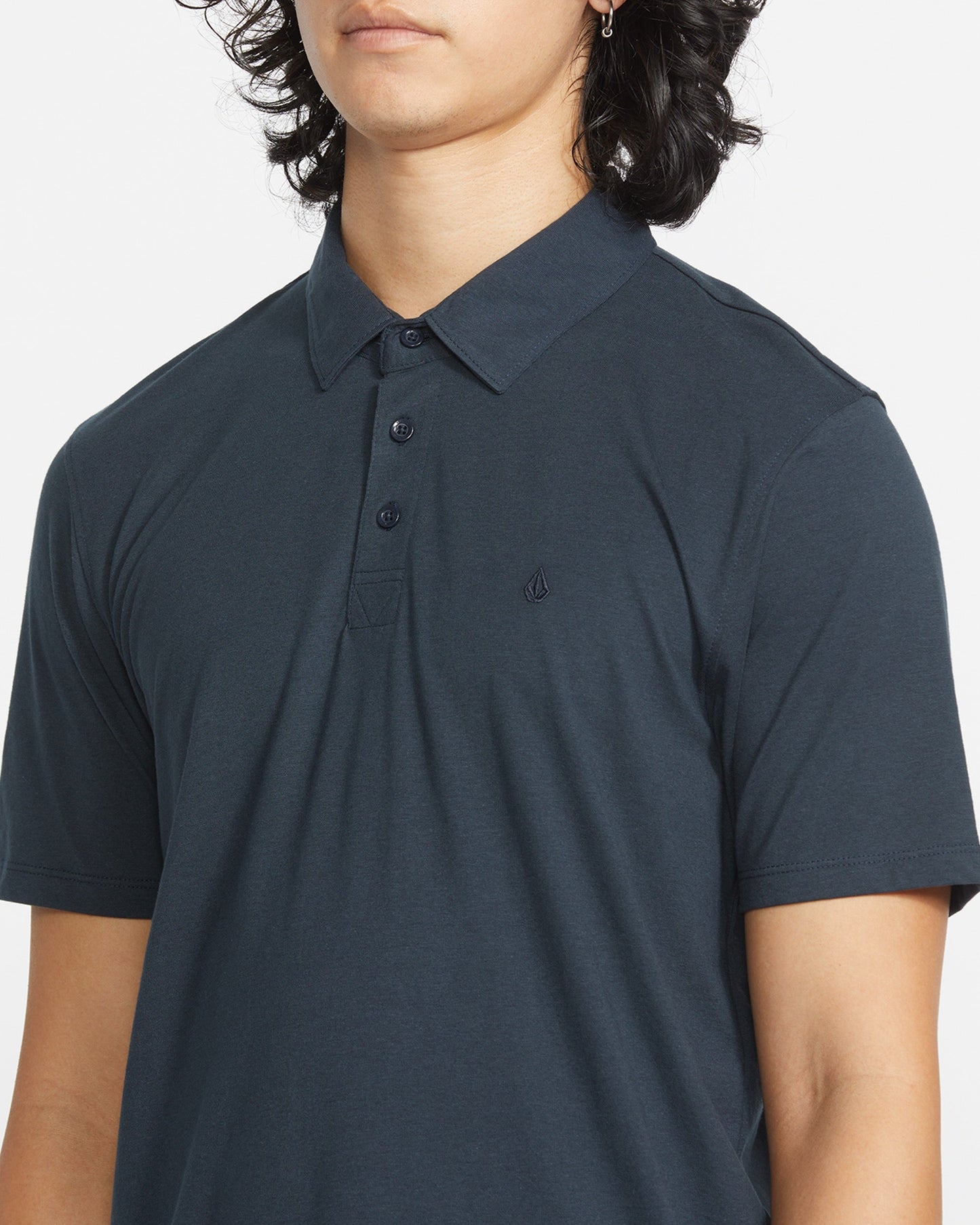 A person with shoulder-length dark hair wears the Volcom Men's Wowzer Polo Short Sleeve Shirt in dark blue, featuring a small chest logo. The modern fit polo is made from a comfortable cotton-poly blend, shown cropped at nose and mid-arm.