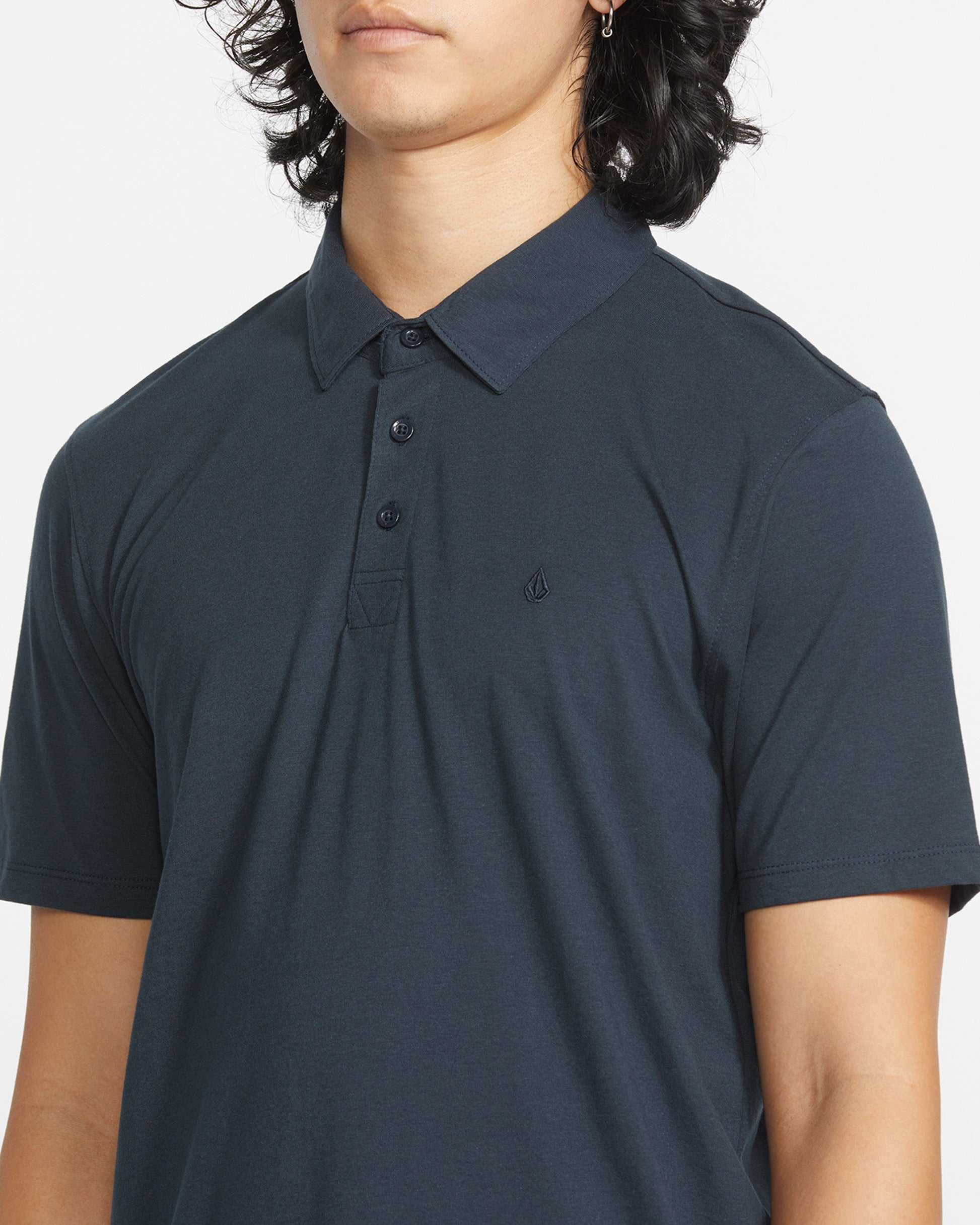 A person with shoulder-length dark hair wears the Volcom Men's Wowzer Polo Short Sleeve Shirt in dark blue, featuring a small chest logo. The modern fit polo is made from a comfortable cotton-poly blend, shown cropped at nose and mid-arm.