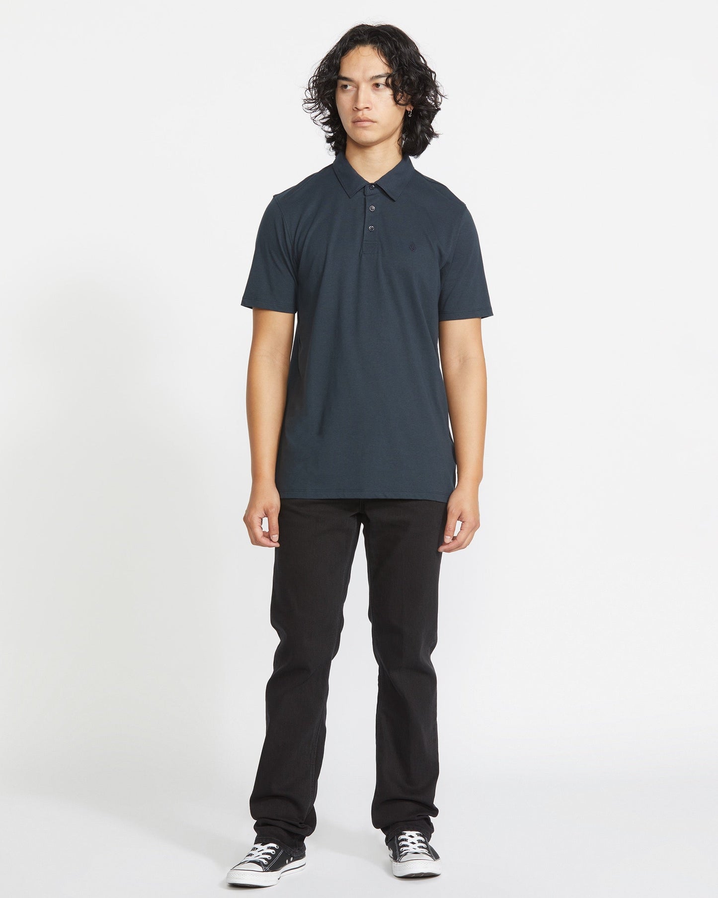 A person with curly dark hair stands against a plain white background, wearing a Volcom Men's Wowzer Polo Short Sleeve Shirt, black pants, and black-and-white sneakers, facing forward with a neutral expression.