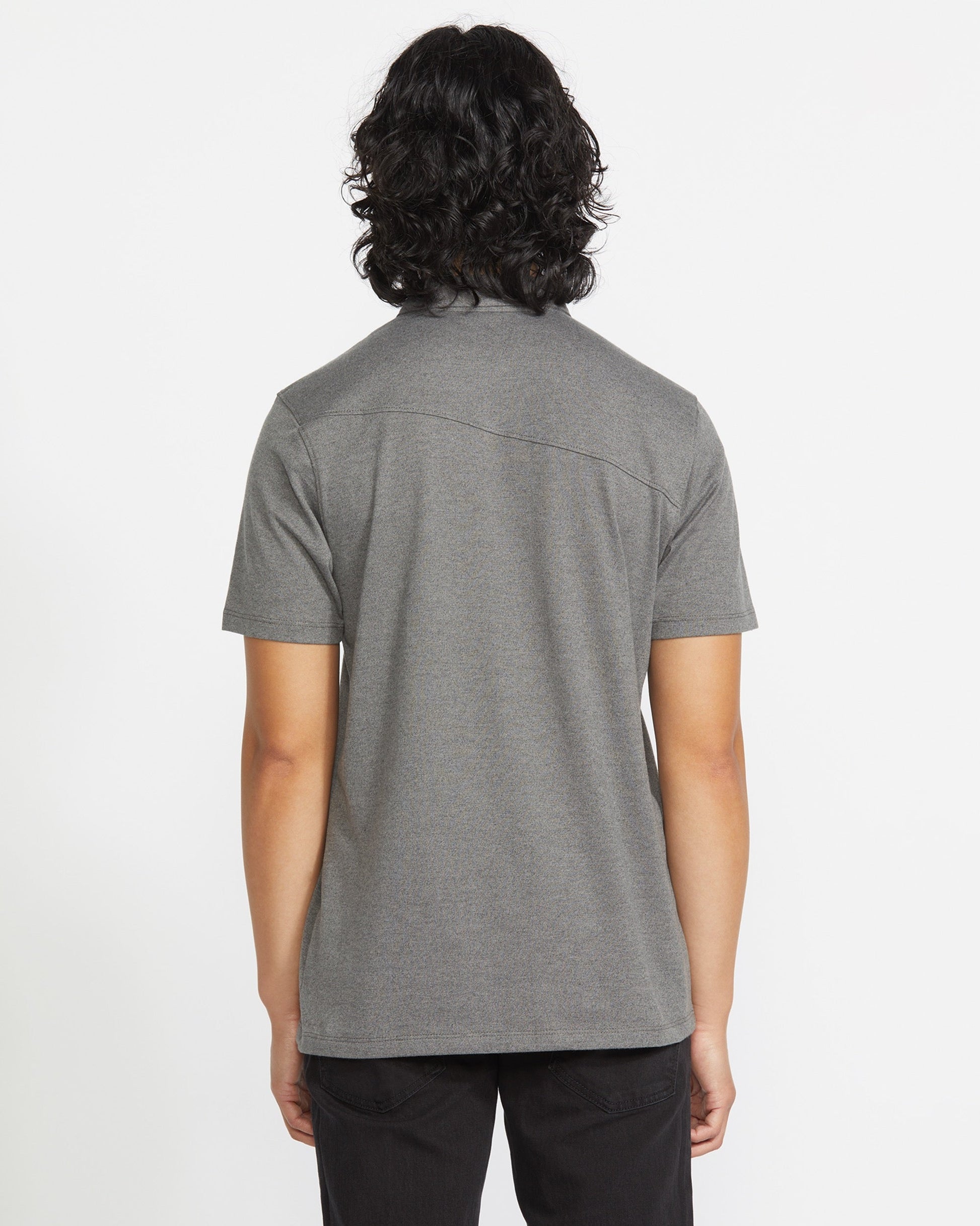 A person with medium-length, wavy dark hair stands with their back to the camera, wearing a Volcom Men's Wowzer Polo Short Sleeve Shirt and black pants against a plain white background.
