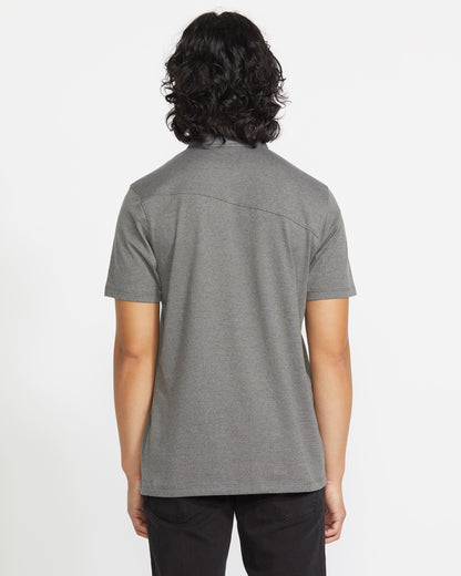 A person with medium-length, wavy dark hair stands with their back to the camera, wearing a Volcom Men's Wowzer Polo Short Sleeve Shirt and black pants against a plain white background.