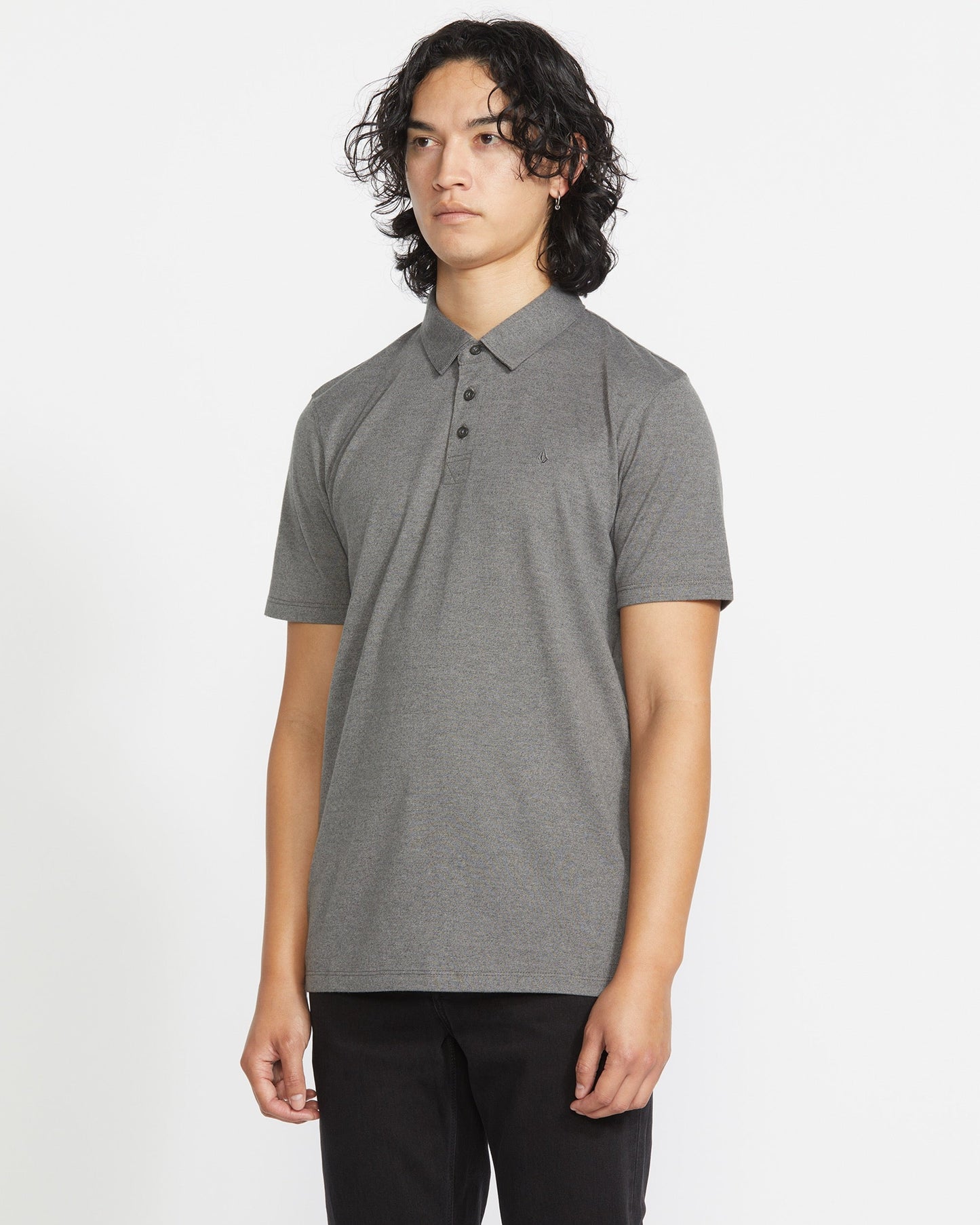 A person with wavy, shoulder-length hair wears the Volcom Men's Wowzer Polo Short Sleeve Shirt in gray and black pants, standing against a plain white background and looking forward with a neutral expression.