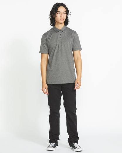 A man with medium-length curly hair wears the Volcom Men's Wowzer Polo Short Sleeve Shirt, featuring chest Stone embroidery, styled with black pants and black sneakers, against a plain white background.