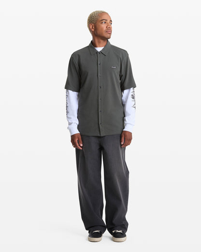 A person with short blonde hair wears a Volcom Men's Chavo Button Down Short Sleeve over a long-sleeve white printed top, loose dark jeans, and black sneakers, standing against a plain white background.
