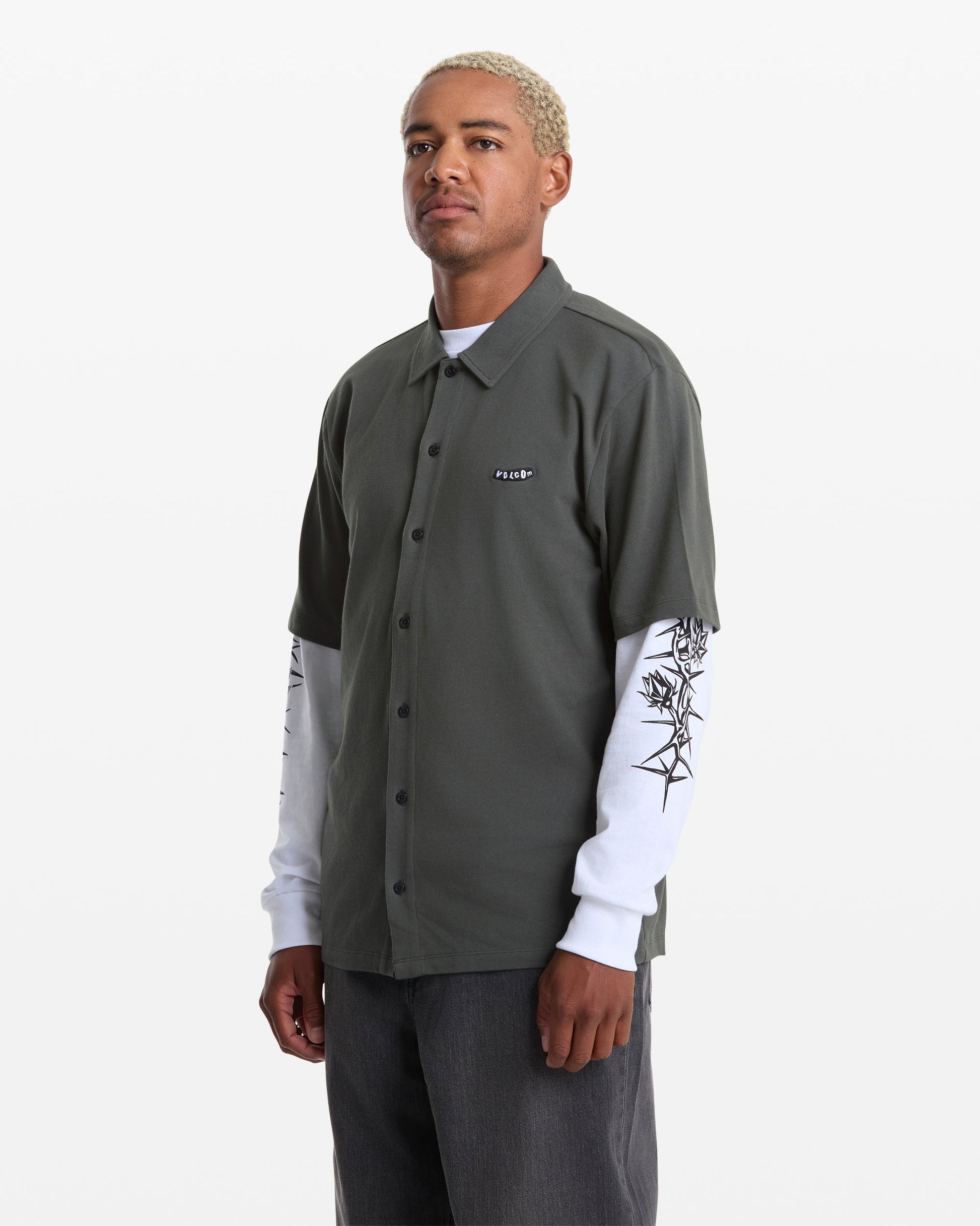A person with short, bleached hair wears a dark green Volcom Men's Chavo Button Down Short Sleeve shirt over a white long-sleeve with black sleeve designs and dark pants, standing against a plain white background.