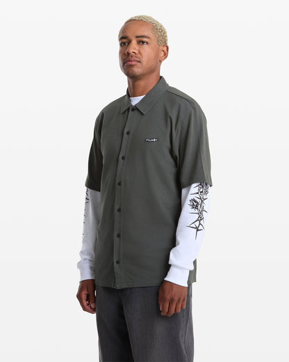 A person with short, bleached hair wears a dark green Volcom Men's Chavo Button Down Short Sleeve shirt over a white long-sleeve with black sleeve designs and dark pants, standing against a plain white background.