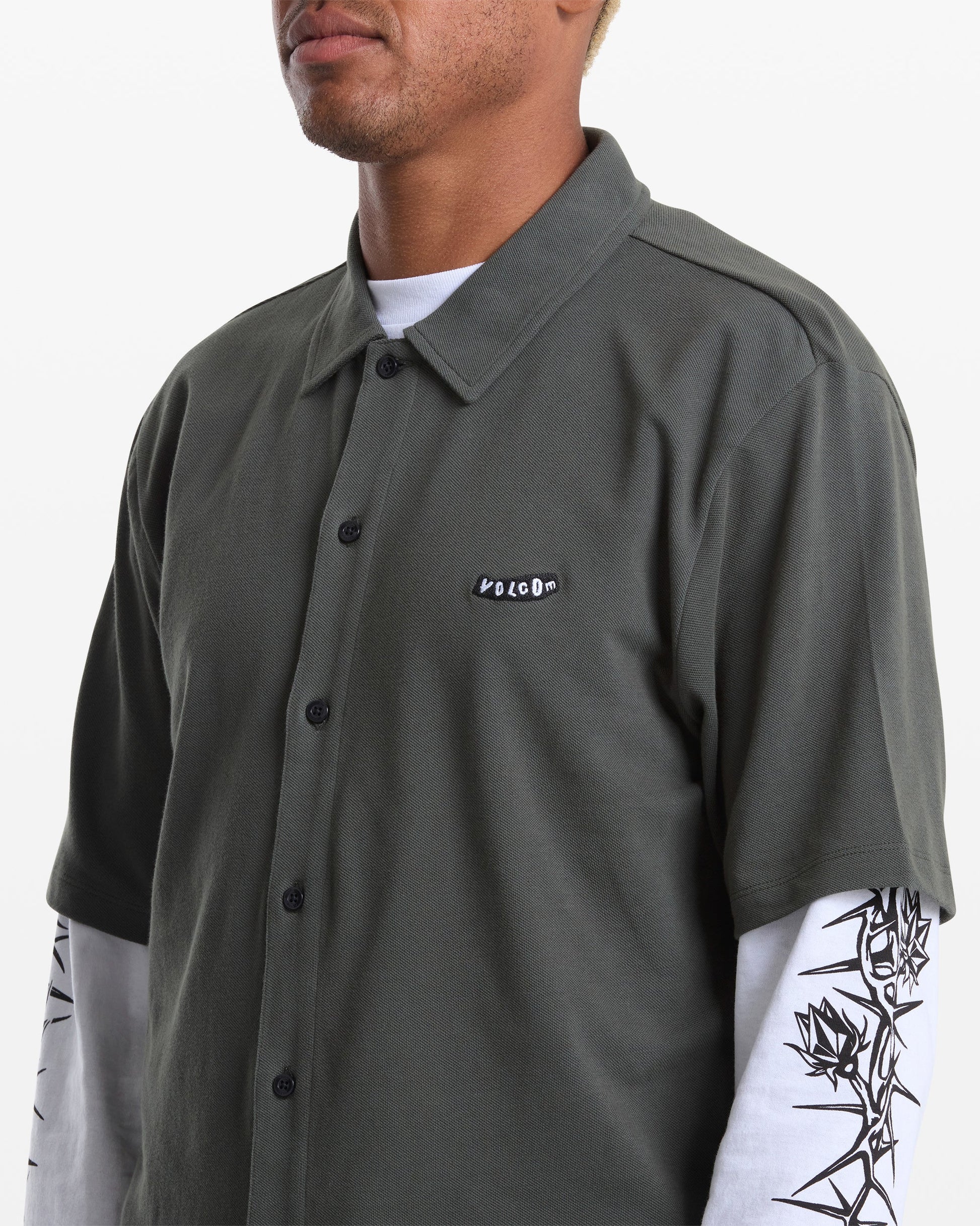 A person wears the Volcom Men's Chavo Button Down Short Sleeve shirt in dark green cotton pique, featuring a logo patch, layered over a white long-sleeve with black graphic artwork on the sleeves.