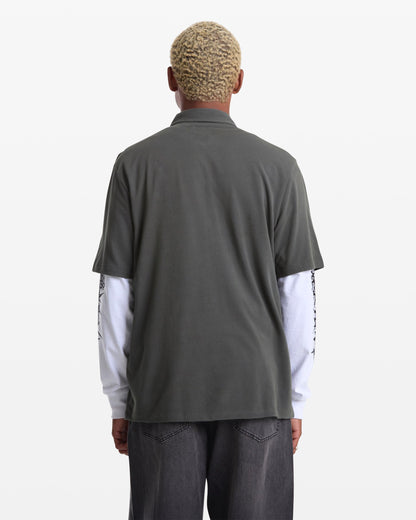 A person with short, curly blond hair is seen from behind wearing the Volcom Men's Chavo Button Down Short Sleeve shirt in dark gray textured cotton pique over a white long-sleeve shirt and dark gray jeans, against a plain white background.