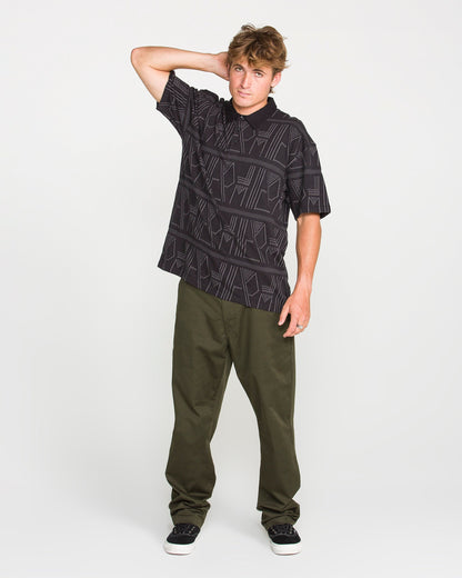 A young man wearing the Volcom Men's Stone Optix Polo—black with a distinctive pattern, boxy fit, and crafted from yarn-dyed jacquard—and olive green pants stands with one hand behind his head and the other by his side, posing against a plain white background.