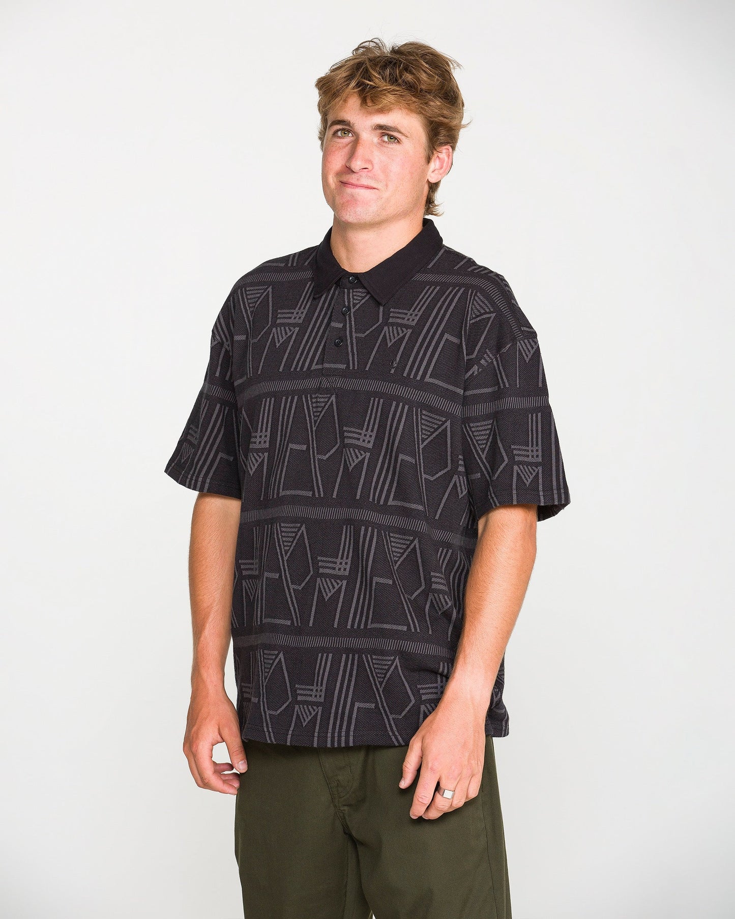 A person with short brown hair wearing the Volcom Men's Stone Optix Polo in a black, boxy fit crafted from yarn-dyed jacquard, paired with olive green pants, stands against a plain white background and looks at the camera with a neutral expression.