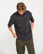 A person with light brown hair wearing the Volcom Men's Stone Optix Polo in black, which features a boxy fit and a yarn-dyed jacquard pattern, paired with olive green pants, looks down while adjusting their shirt against a plain white background.
