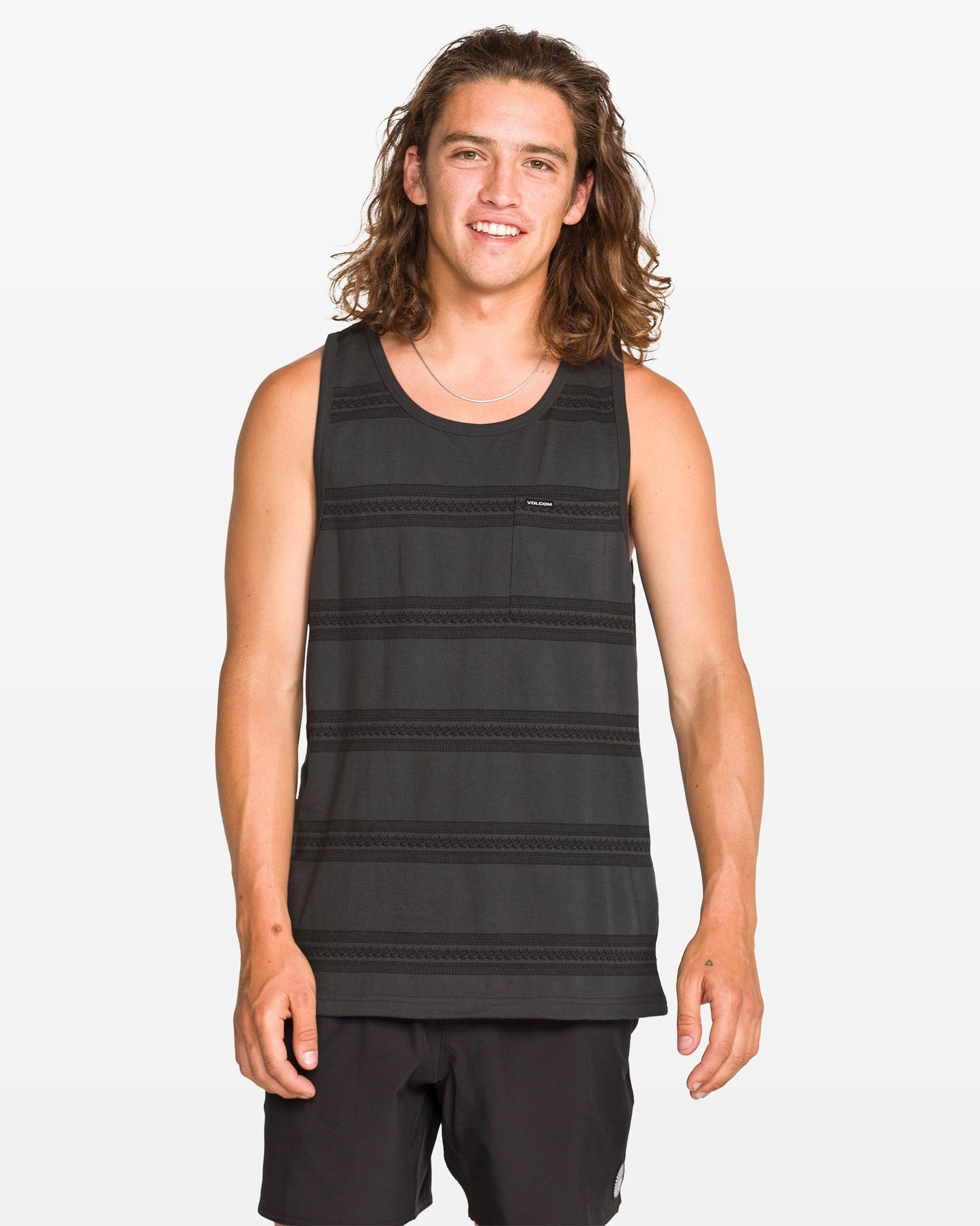 A young man with long brown hair stands and smiles at the camera against a plain white background, wearing a modern fit Men's Yewster Tank by Volcom in black and gray jacquard stripes paired with black shorts.