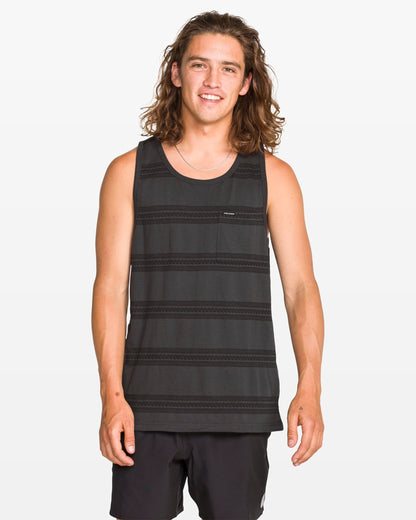 A young man with long brown hair stands and smiles at the camera against a plain white background, wearing a modern fit Men's Yewster Tank by Volcom in black and gray jacquard stripes paired with black shorts.