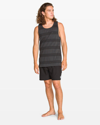 A man with long brown hair stands barefoot in Volcom's Men's Yewster Tank and black shorts, looking to his left against a plain white background.