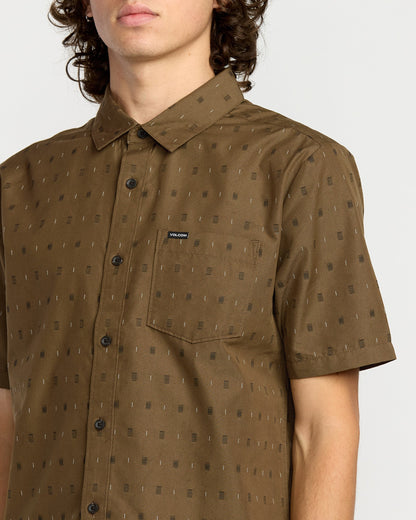 A man wears the Volcom Men's Bankstone Short Sleeve Shirt in brown, featuring a classic fit, subtle geometric pattern, chest pocket with a Volcom label, and short sleeves. His face is partially out of frame.