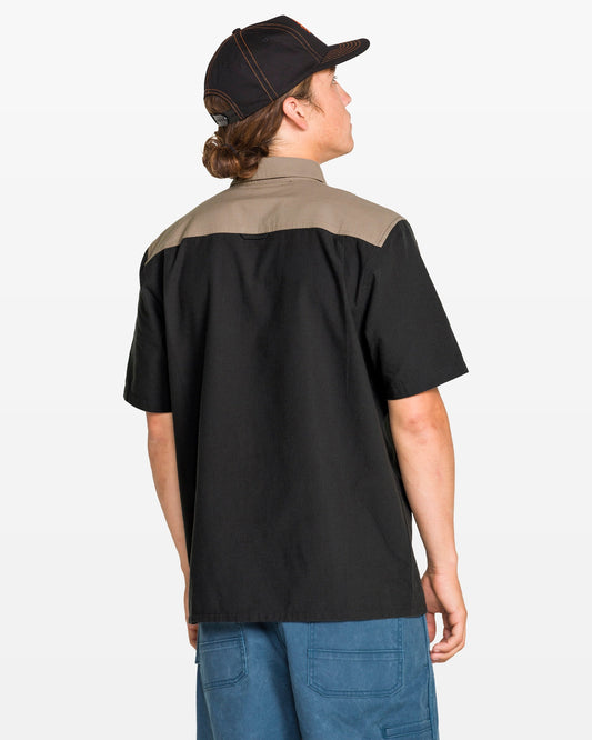 A person with long hair in a ponytail, wearing the Volcom Men's Slab City Short Sleeve Tee, blue pants, and a black cap stands with their back to the camera against a plain background.