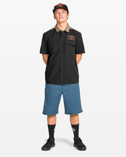 A young man stands facing forward in a classic fit black Volcom Men's Slab City Short Sleeve Tee, blue knee-length shorts, black socks, black shoes, and a black cap, set against a plain white background.