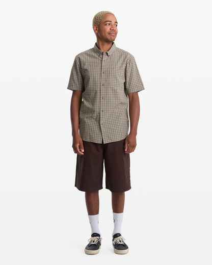 A person with short blonde hair wears the Volcom Men's Brewster Short Sleeve shirt, brown knee-length shorts, white crew socks, and black sneakers, standing against a plain white background.