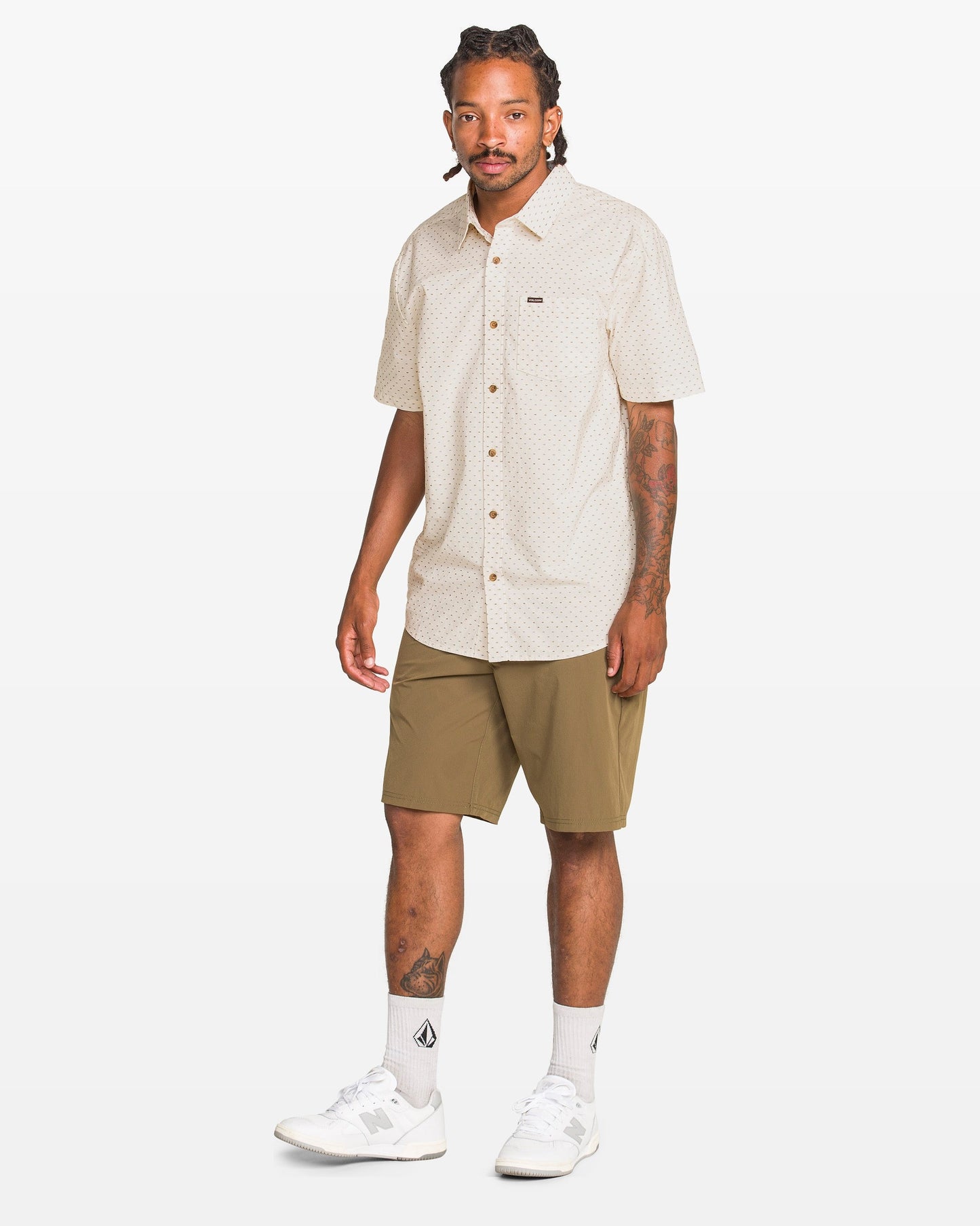 A man with braided hair wears the Volcom Men's Bankstone II Short Sleeve Tee, olive green shorts, white socks, and white sneakers as he stands against a plain white background.