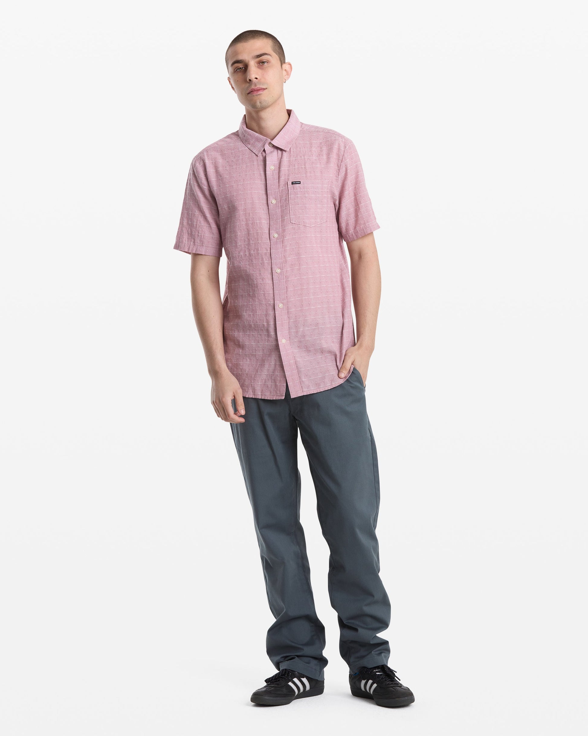 A man with short hair and a neutral expression stands against a white background wearing the Volcom Men's Bankstone II Short Sleeve shirt, blue-gray pants, and black Adidas sneakers with white stripes, one hand in his pocket.