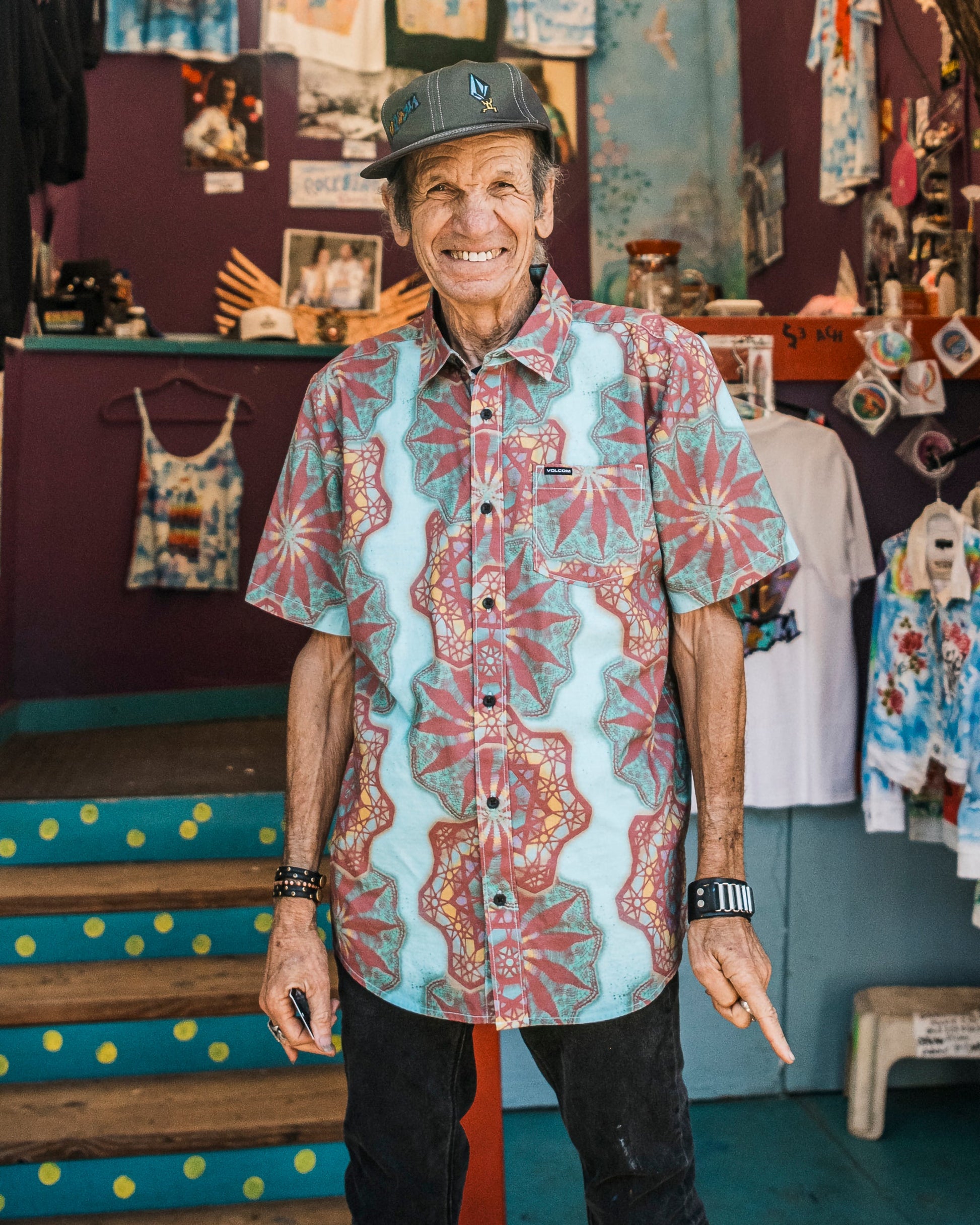 A man in a Volcom Men's Star Shields Featured Artist Short Sleeve Shirt in Castlerock and a cap smiles warmly while standing in a vibrant, art-filled store decorated with t-shirts and crafts.