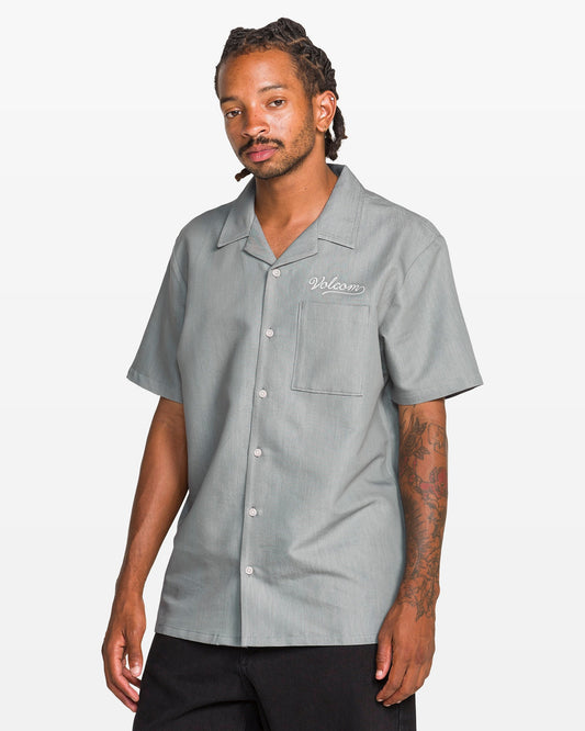 A man with tattoos on his right arm and braided hair wears the Volcom Men's Otis Stone Short Sleeve Tee, a boxy fit, textured light gray shirt with "Welcome" embroidered on the chest pocket. He stands against a plain white background.