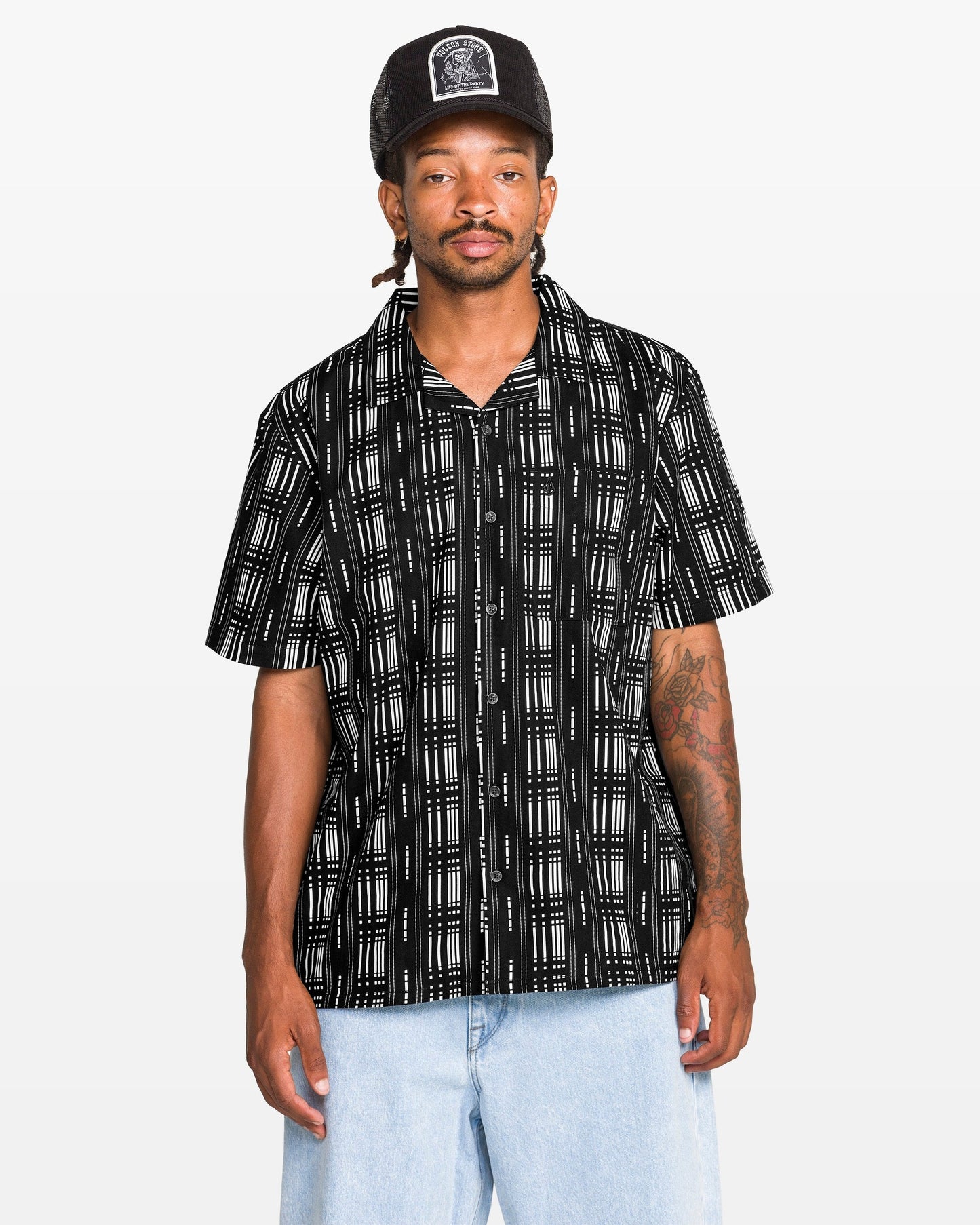 A man stands against a plain white background wearing the Volcom Men's Macros Short Sleeve Tee, styled with light blue jeans and a black cap.
