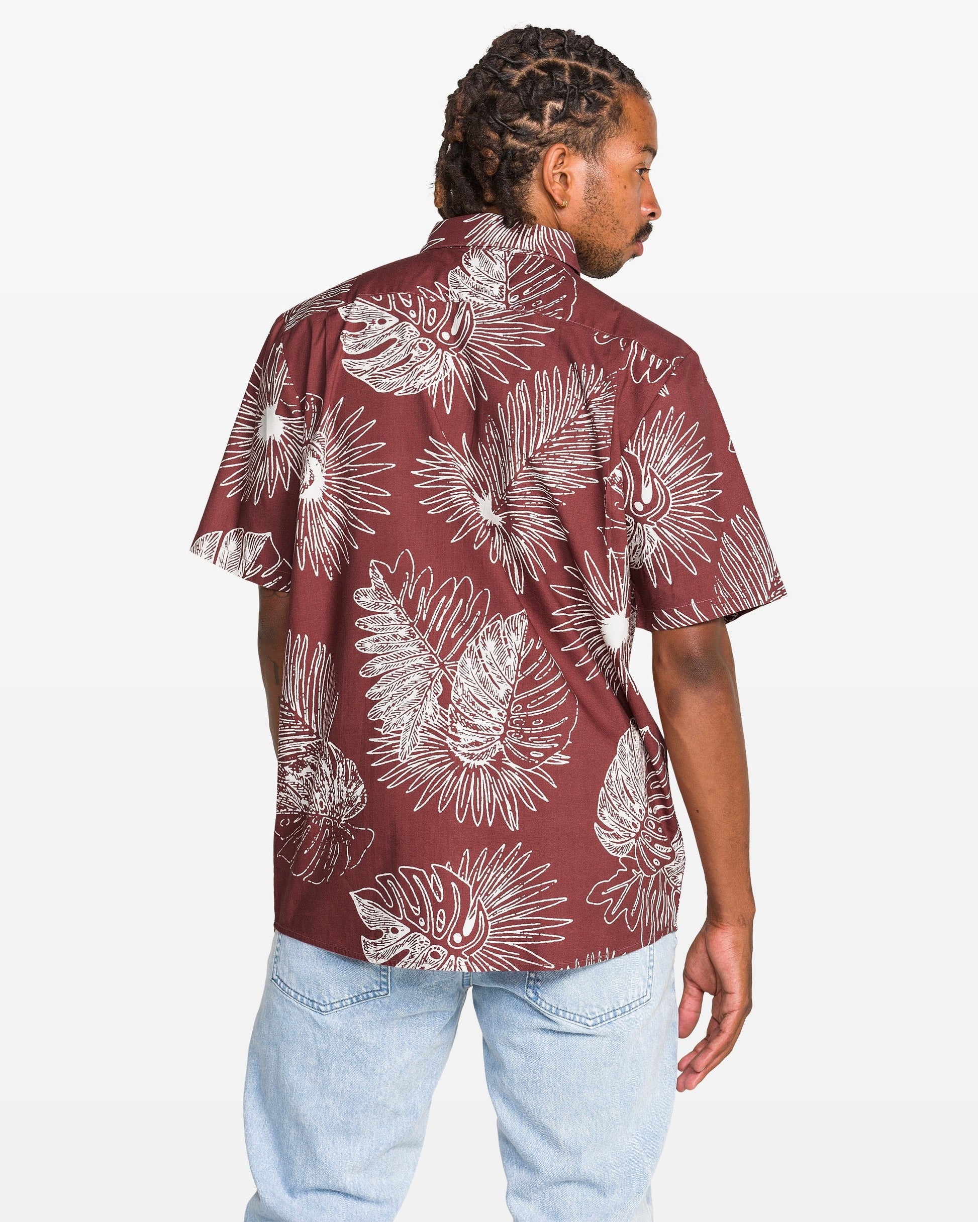A man with braided hair wears the Volcom Men's Monsterious Short Sleeve Shirt—a cotton viscose blend with white tropical leaf print on maroon—and light blue jeans. He stands with his back to the camera, looking over his shoulder.