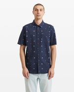 A person stands facing forward against a plain white background, wearing the Volcom Men's Scaler Stone Woven Short Sleeve Shirt—an allover circular dot print shirt in navy blue—with light-colored pants.