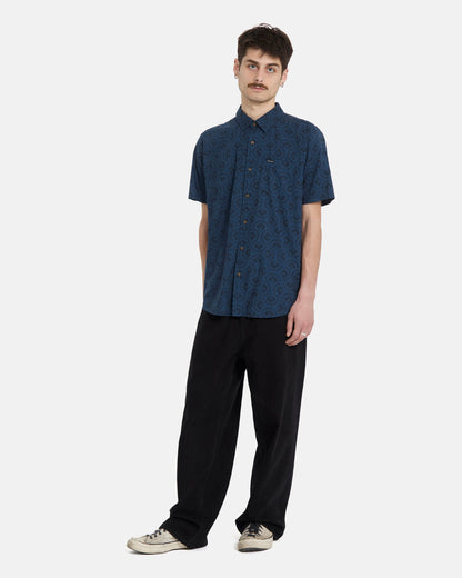 A man with short brown hair and a mustache wears the Volcom Men's Scaler Stone Woven Short Sleeve Shirt in midnight blue, paired with loose black pants and white sneakers, standing against a plain white background.