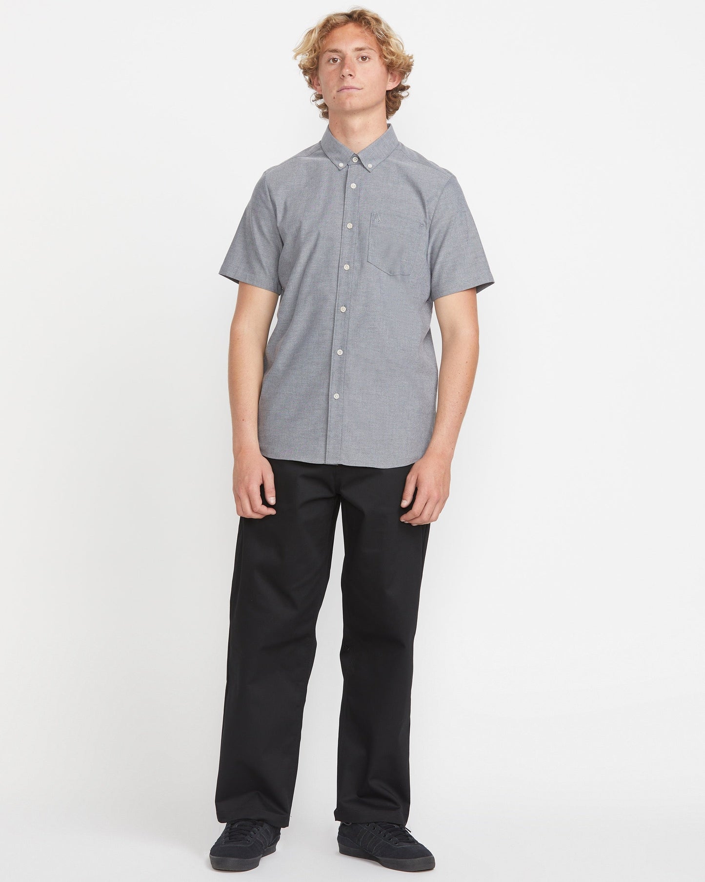 A person with curly blonde hair stands facing forward against a white background, wearing Volcom's Men's Everett Oxford Short Sleeve Tee in gray and black pants.