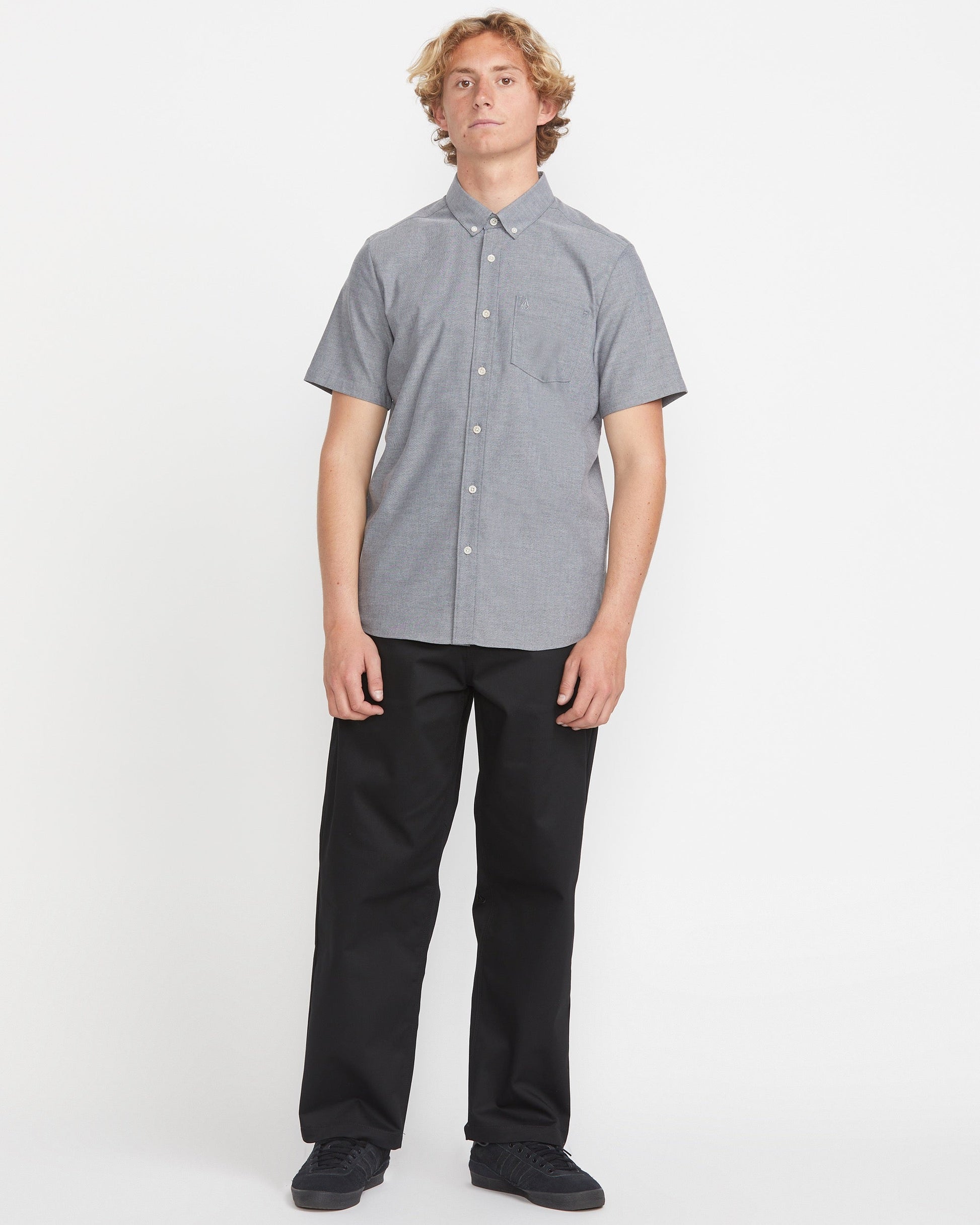 A person with curly blonde hair stands facing forward against a white background, wearing Volcom's Men's Everett Oxford Short Sleeve Tee in gray and black pants.