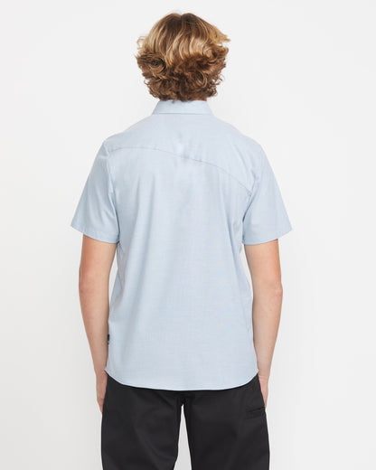 A person with wavy, light brown hair stands facing away from the camera, wearing the Volcom Men's Everett Oxford Short Sleeve in light blue and black pants against a plain white background.