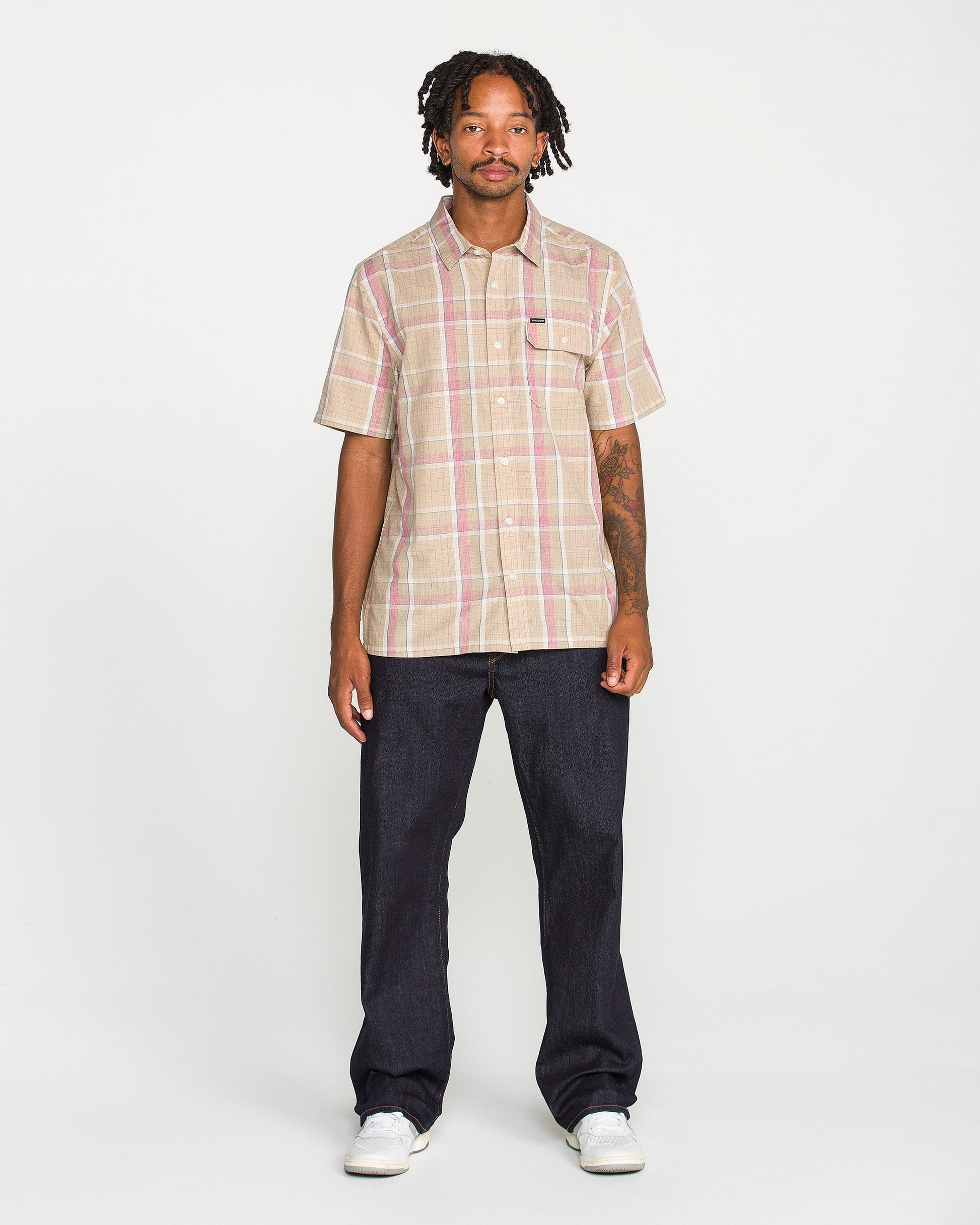 A man stands facing forward, wearing the Volcom Men's Ledges Short Sleeve Shirt in a classic-fit yarn-dyed plaid, paired with dark jeans and white sneakers against a plain white background.