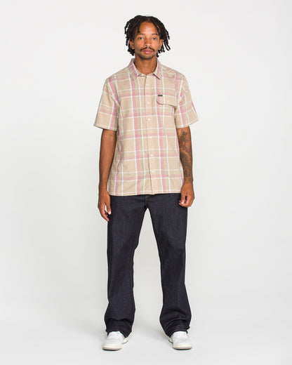 A man stands facing forward, wearing the Volcom Men's Ledges Short Sleeve Shirt in a classic-fit yarn-dyed plaid, paired with dark jeans and white sneakers against a plain white background.