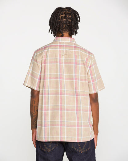 A person with short, twisted hair and visible tattoos on their left arm stands with their back to the camera, wearing the Volcom Men's Ledges Short Sleeve Shirt—a classic-fit, beige yarn-dyed plaid shirt with pink and white stripes—paired with dark jeans.