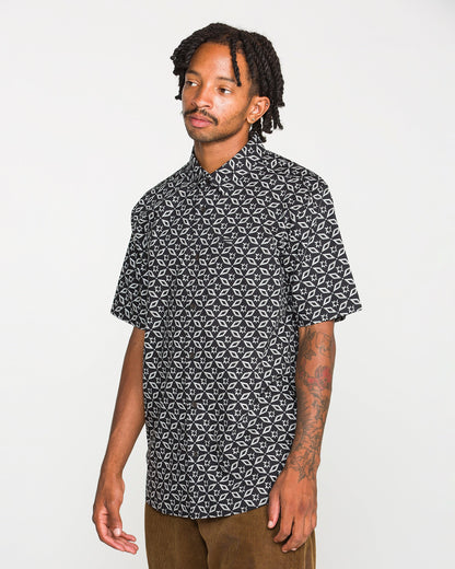 A man with short dreadlocks and tattoos on his arm wears the Volcom Men's Scaler Stone Short Sleeve Shirt and brown pants, standing against a plain white background.