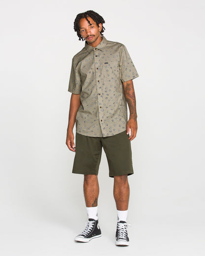 A man stands against a white background, wearing the Volcom Men's Scaler Stone Short Sleeve Shirt featuring an eye-catching pattern, paired with olive green knee-length shorts, white socks, and black high-top sneakers. He has short dreadlocks and visible tattoos on one arm.