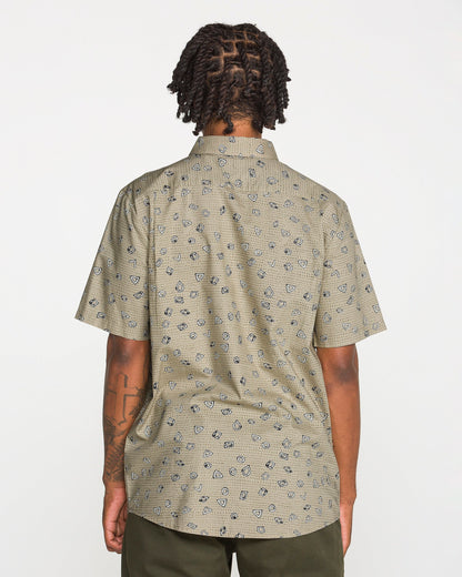 A person with short, twisted hair is photographed from the back against a plain white background wearing Volcom's Men's Scaler Stone Short Sleeve Shirt, featuring a classic fit and beige pattern, paired with olive green pants.