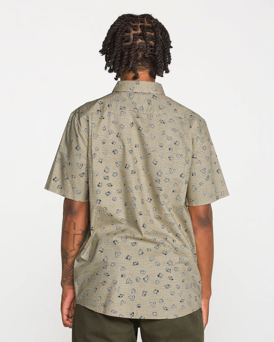 A person with short, twisted hair is photographed from the back against a plain white background wearing Volcom's Men's Scaler Stone Short Sleeve Shirt, featuring a classic fit and beige pattern, paired with olive green pants.
