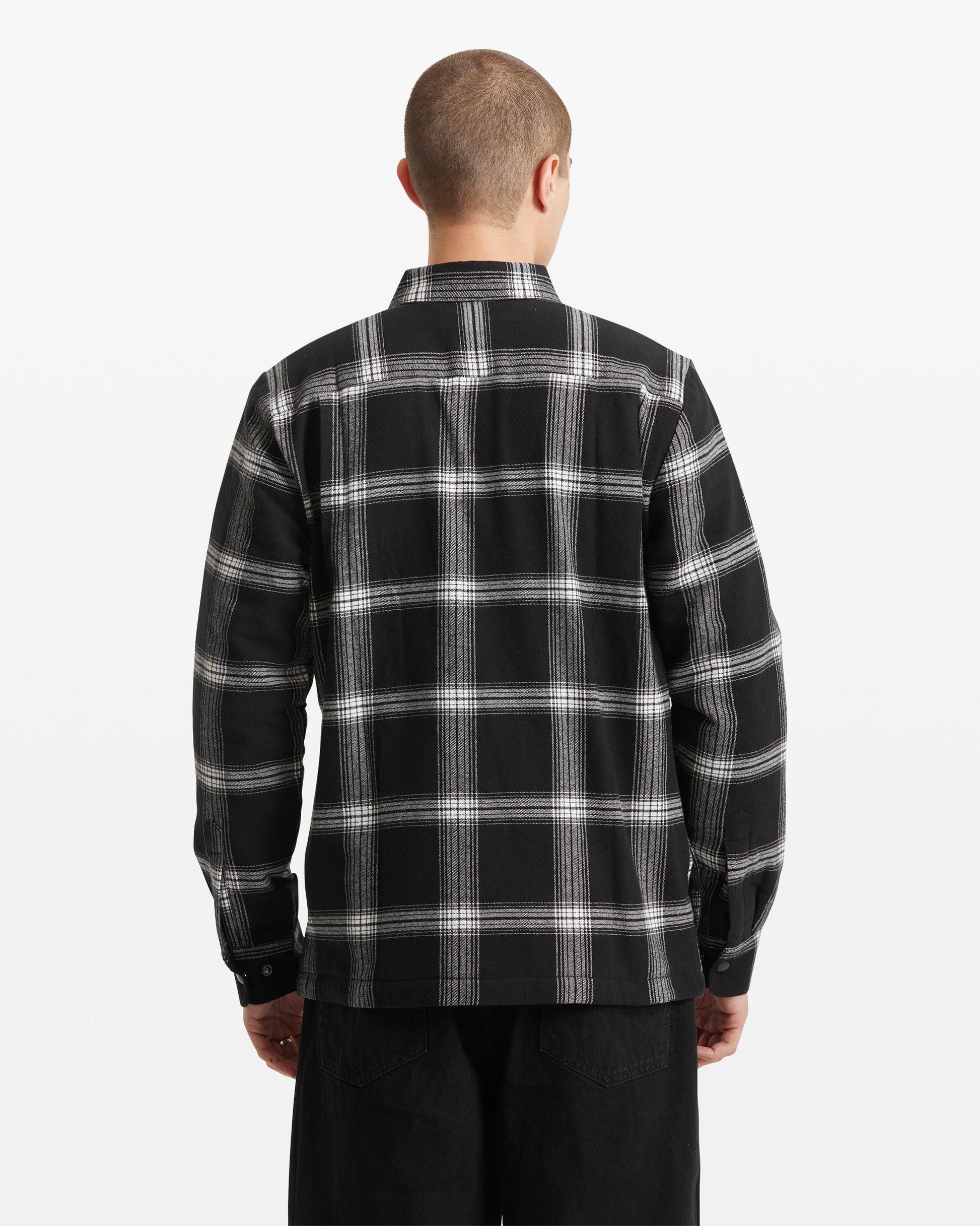 A person with short hair is shown from the back against a plain white background, wearing the Volcom Men's Wilderstone Long Sleeve in a black and white plaid pattern with black pants.