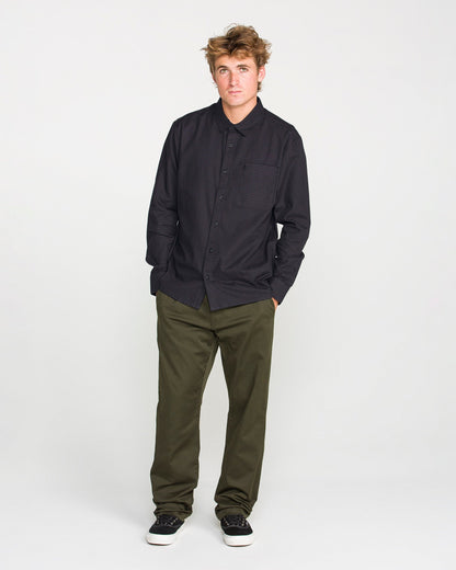 A young man with tousled hair stands against a plain white background, wearing the Volcom Men's Glasnow Long Sleeve Shirt in a dark, boxy fit with a yarn-dyed dobby texture, paired with olive green pants and black sneakers with white soles. His hands are in his pockets as he looks slightly to the side.
