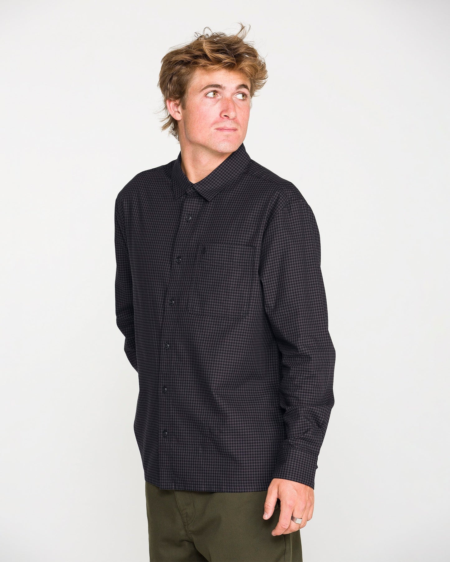 A man with tousled hair wears the Volcom Men's Glasnow Long Sleeve Shirt, a black checkered, yarn-dyed dobby texture button-up, paired with olive green pants as he looks to his left against a plain white background.