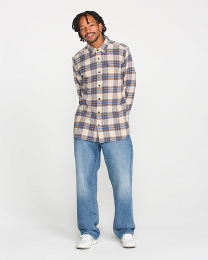 A man wearing the Volcom Men's Caden Plaid Long Sleeve Shirt in a classic beige and blue pattern, paired with loose blue jeans and white sneakers, stands against a plain white background, smiling with his hands in his pockets.