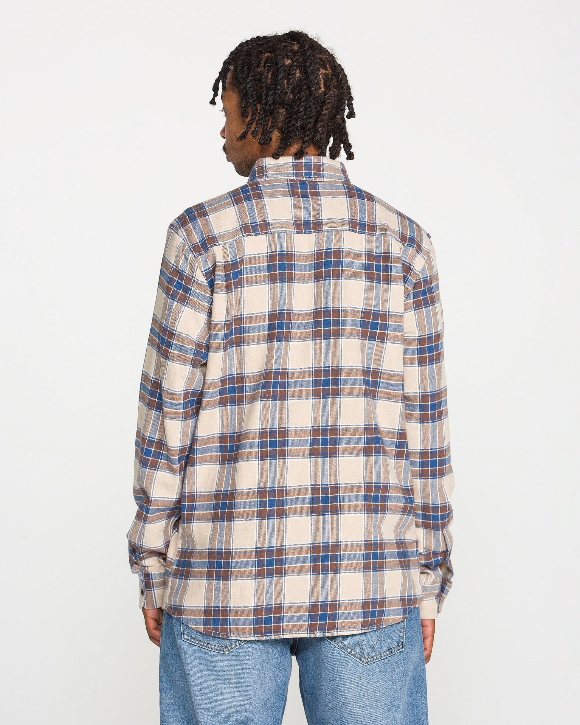 A person with short dreadlocks is shown from the back, wearing the Volcom Men's Caden Plaid Long Sleeve Shirt in beige, blue, and brown, paired with light blue jeans against a plain white background.