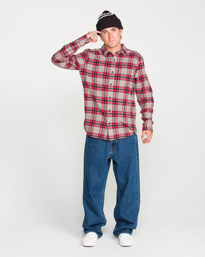 A person wearing a black beanie, blue jeans, white sneakers, and the Volcom Men's Caden Plaid Long Sleeve Shirt in red stands against a plain white background, touching the brim of their beanie with one hand.